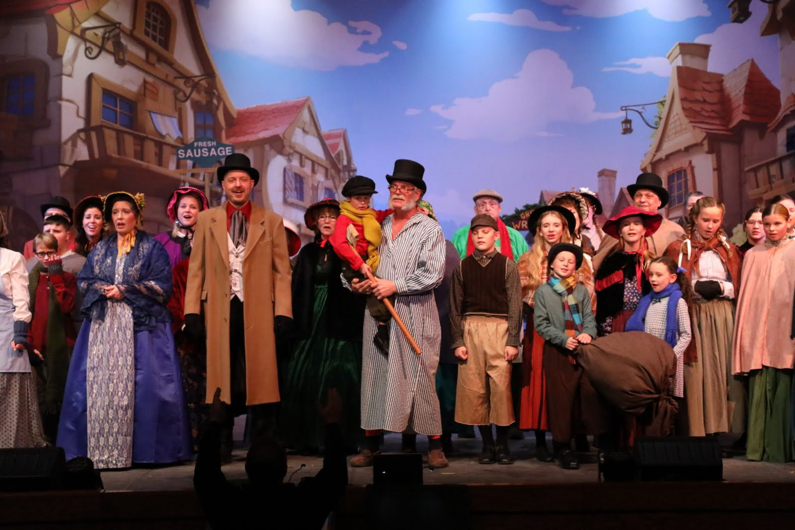 Men, women and children in Dickens costumes in front of a town square drop with Scroofe holding Tiny Tim.